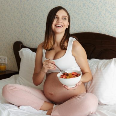 Portrait of a laughing young pregnant woman having healthy breakfast while sitting on bed with a bowl and spoon
