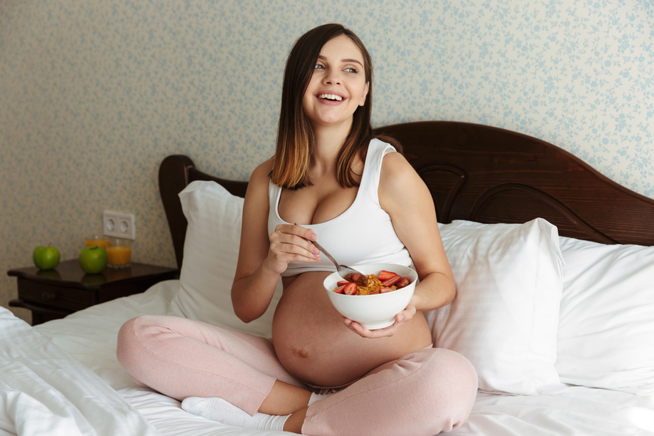 Portrait of a laughing young pregnant woman having healthy breakfast while sitting on bed with a bowl and spoon