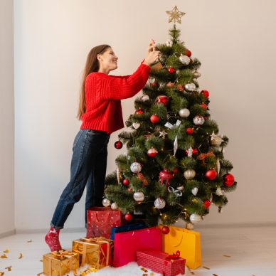 smiling excited pretty woman in red sweater standing at home decorating Christmas tree surrounded with presents and gift boxes