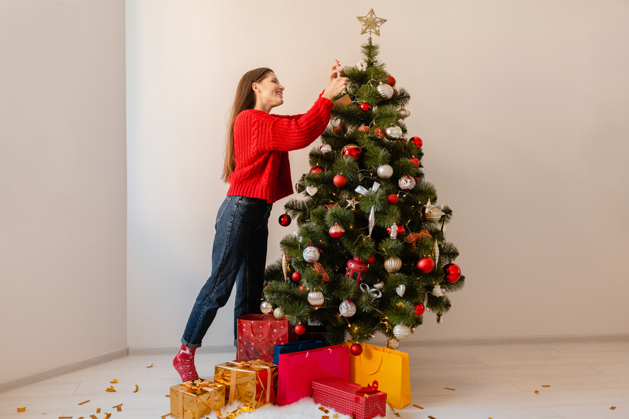 smiling excited pretty woman in red sweater standing at home decorating Christmas tree surrounded with presents and gift boxes