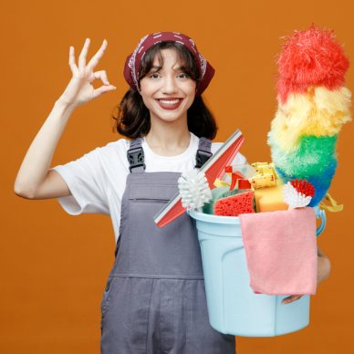 smiling young female cleaner wearing uniform and bandana holding bucket of cleaning tools looking at camera showing ok sign isolated on orange background