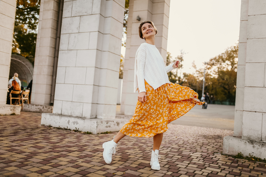 young pretty happy smiling woman in yellow printed dress and knitted white sweater on sunny autumn day having fun in street wearing stylish outfit and white boots