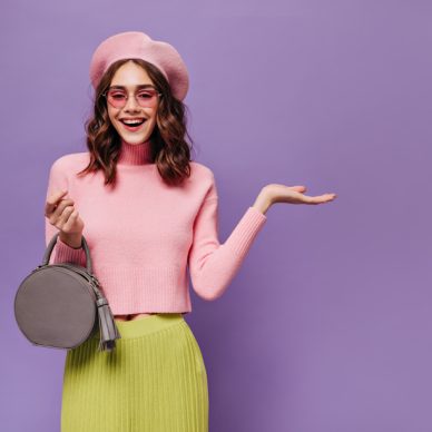 Joyful Parisian woman in beret and sunglasses points at place for text on purple background. Attractive girl in pink sweater and green skirt smiles.