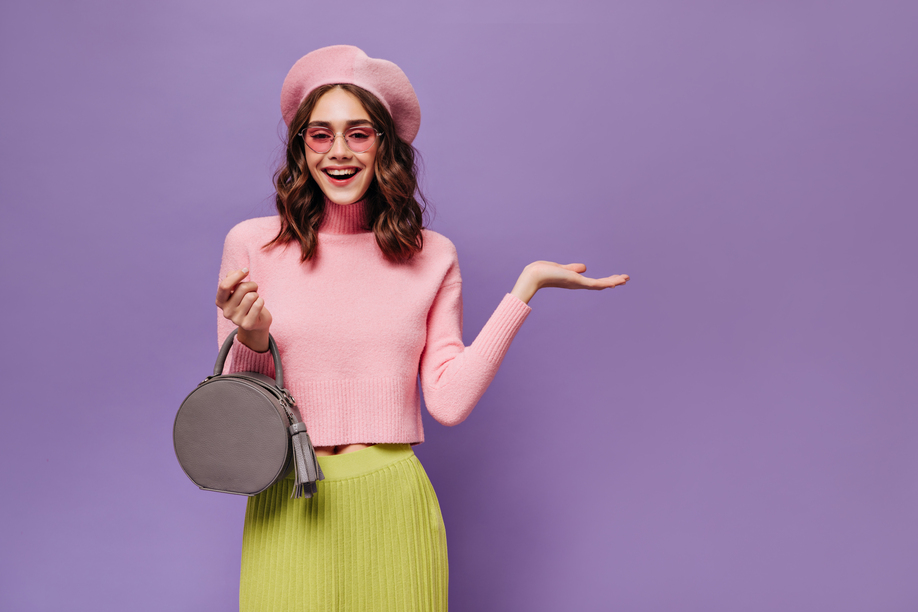 Joyful Parisian woman in beret and sunglasses points at place for text on purple background. Attractive girl in pink sweater and green skirt smiles.