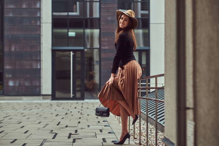 Smiling fashion elegant woman wearing a black jacket, brown hat and skirt with a handbag clutch posing while leaning on a steel railing on a European city center.