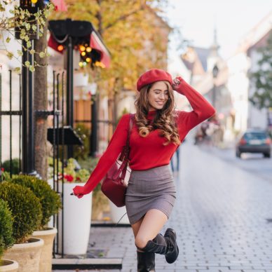 Joyful young woman in red beret dancing on pavement with charming smile. Outdoor portrait of graceful white girl in elegant outfit fooling around on the street.