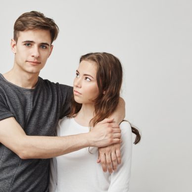 Portrait of couple hugging near the wall. Girlfriend thinks that he continues talk with his ex so she looks suspicious. Boyfriend trying to act like nothing happened and avoids looking in her eyes. Relationship concept