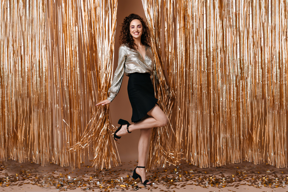 Positive lady in black skirt and shiny top coquettishly raising leg on gold background