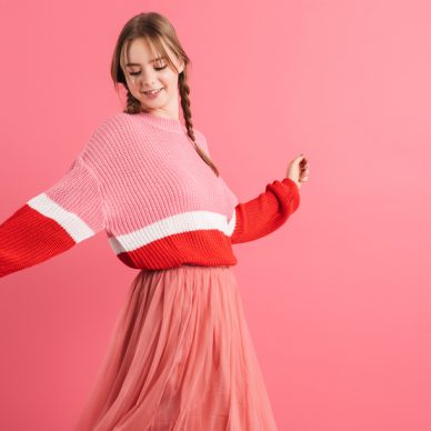 Young pretty smiling girl with two braids in sweater and long skirt happily dancing spending time over pink background