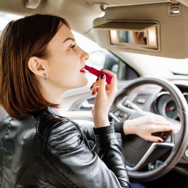 Businesswoman looking in rear view mirror and making up her lips with red lipstick in car