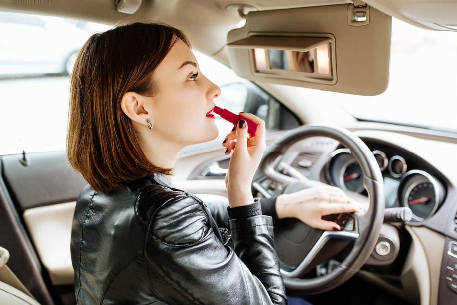 Businesswoman looking in rear view mirror and making up her lips with red lipstick in car