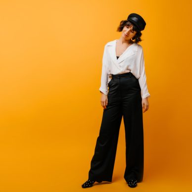 Full-length portrait of stylish woman in black pants and hat. Amazing short-haired girl in white shirt standing on yellow background.