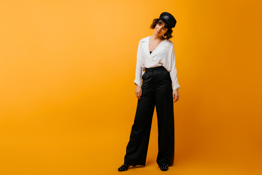Full-length portrait of stylish woman in black pants and hat. Amazing short-haired girl in white shirt standing on yellow background.