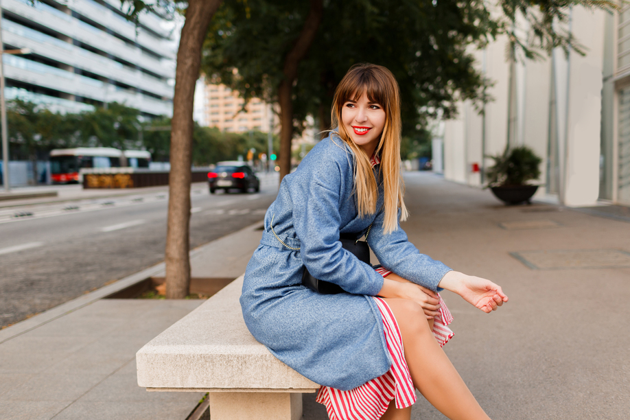 Outdoor portrait of  stylish pretty  happy  woman in blue coat sitting on bench on urban background.