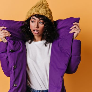 Sensual young woman in purple down jacket looking away. Studio shot of cute mixed race girl in knitted hat.