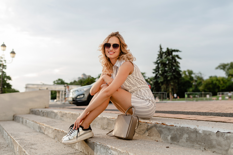 young attractive stylish blonde woman sitting in city street in summer fashion style dress wearing sunglasses, purse, silvers sneakers footwear style