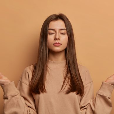 Peaceful calm relaxed young woman meditates indoor, keeps hands in zen gesture, closes eyes, practices yoga, finds peace inside of soul, reaches nirvana, dressed casually, poses indoor over beige wall