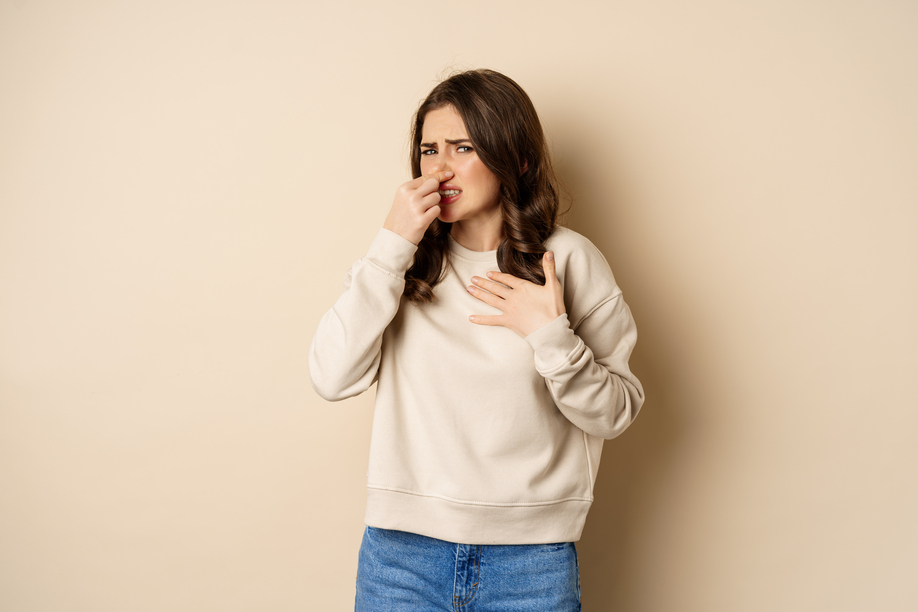 Woman expressing disgust of bad disgusting smell, close nose and grimaing from dislike, standing over beige background.
