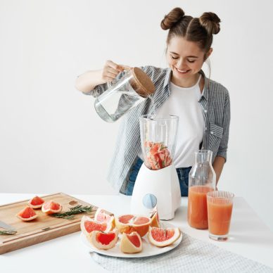 Attractive young girl smiling adding water in blender with grapefruit pieces and rosemary. Healthy diet food nutrition. Copy space. White wall background.