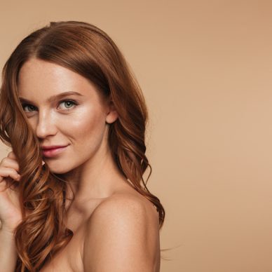 Beauty portrait of mystery smiling ginger woman with long hair posing sideways and looking at the camera over cream background