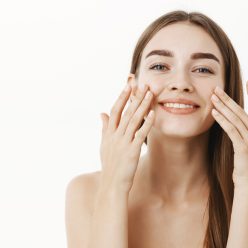 Waist-up shot of charming relaxed and gentle young woman making cosmetological procedure applying facial cream on face with fingers and smiling broadly feeling perfect, taking care of skin