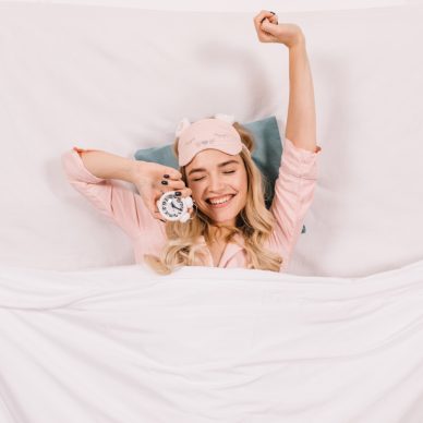 Cheerful girl stretching in bed. Top view of happy blonde woman with clock lying under blanket.