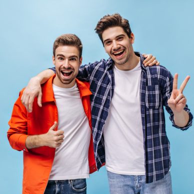 Portrait of charming brunet men in white t-shirts and jeans on isolated. Man in orange jacket show thump up and guy in checkered shirt show peace sign.