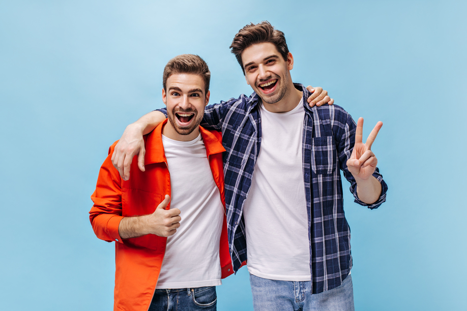 Portrait of charming brunet men in white t-shirts and jeans on isolated. Man in orange jacket show thump up and guy in checkered shirt show peace sign.