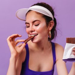 Sweet girl in cap and purple top bites with pleasure milk chocolate. Brunette posing on purple background.