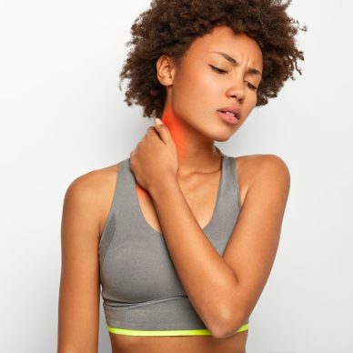 Cropped image of upset curly haired young woman suffers from neck pain, being injured during sport training, wears casual grey top, focused down, isolated over white background. Health problems