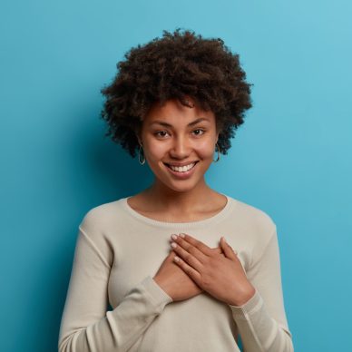 Kind positive African American woman expresses gratitude, has good feeling, presses hands to heart, smiles pleasantly, appreciates someones help, dressed in caual clothes, isolated on blue background