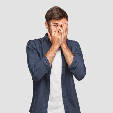 Handsome man with dark hair, bristle, hides face with hands, peeks through fingers, wears fashionable shirt, feels shy, isolated over white background. Curious guy cannot to look at present.