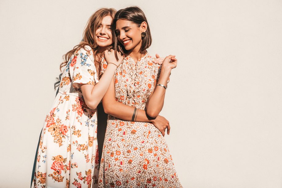 Two young beautiful smiling hipster girls in trendy summer sundress.Sexy carefree women posing near wall on the street background. Positive models having fun and hugging