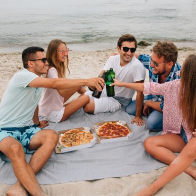 Group of young attractive friends making a toast, drinking beer with pizza while having fun on the beach, near the sea.