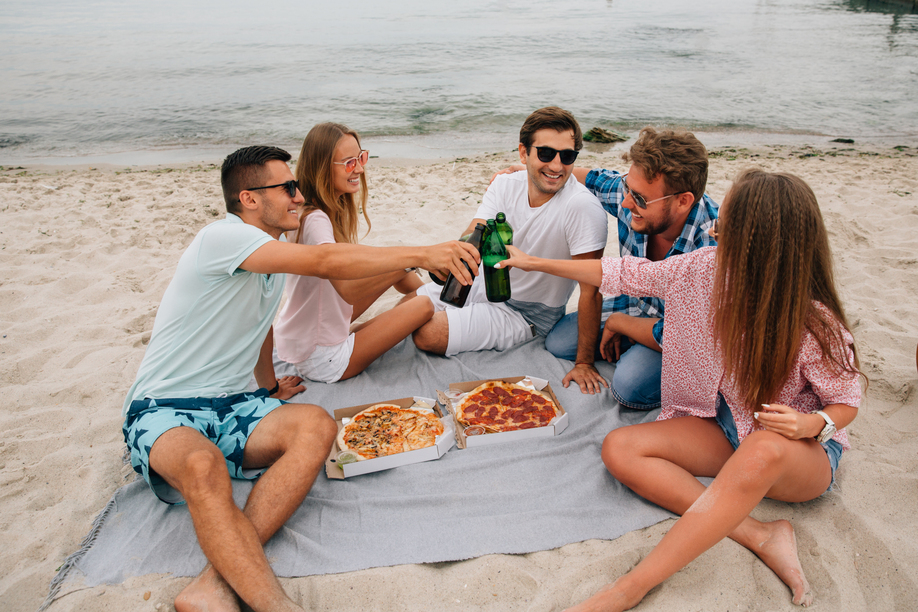 Group of young attractive friends making a toast, drinking beer with pizza while having fun on the beach, near the sea.