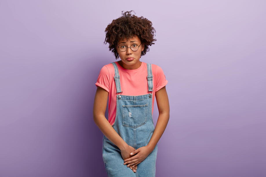 Medical problem, incontience concept. Displeased Afro American woman hods crotch, waits near closed WC, needs toilet, wants to pee, feels discomfort and pain in urinary bladder, wears denim sarafan