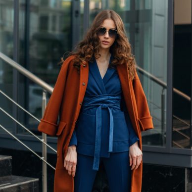 attractive stylish woman with walking in urban city business street dressed in warm brown coat and blue suit, spring autumn trendy fashion street style, wearing sunglasses