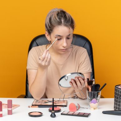 looking at mirror young beautiful girl sits at table with makeup tools applying eyeshadow with makeup brush isolated on orange background