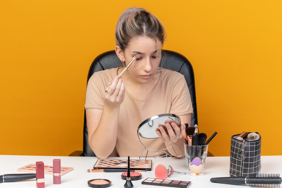 looking at mirror young beautiful girl sits at table with makeup tools applying eyeshadow with makeup brush isolated on orange background