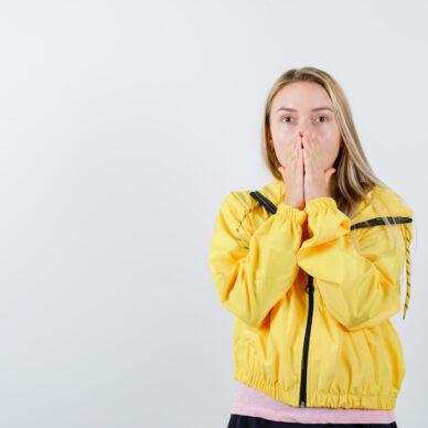 blonde girl holding hands on mouth in yellow jacket and looking scared , front view.