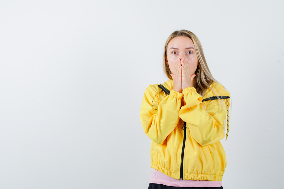 blonde girl holding hands on mouth in yellow jacket and looking scared , front view.