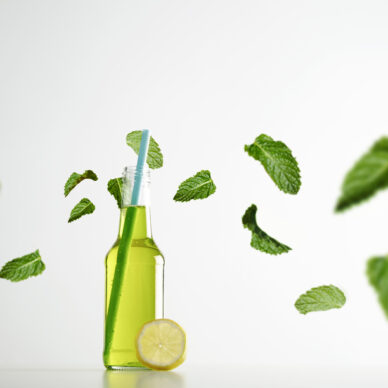 Fresh lime herbal drink inside transparent cocktail opened bottle with blue drinking straw inside, lemon slice near and green mint leaves flying around in air, isolated on white commercial