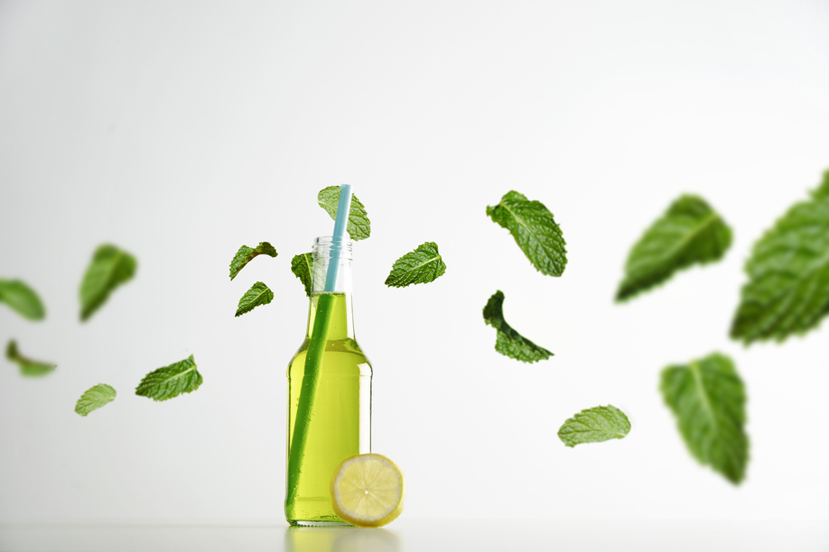 Fresh lime herbal drink inside transparent cocktail opened bottle with blue drinking straw inside, lemon slice near and green mint leaves flying around in air, isolated on white commercial