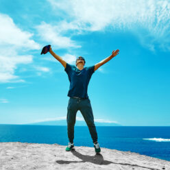 Happy stylish man in casual clothes standing on the cliff of mountain with raised hands to the sun and celebrating success