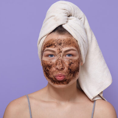portrait of young attractive woman with white towel on her head, posing isolated over lilac background with pouty lips, looks directly at camera, having chocolate scrub or mask for moisturising.