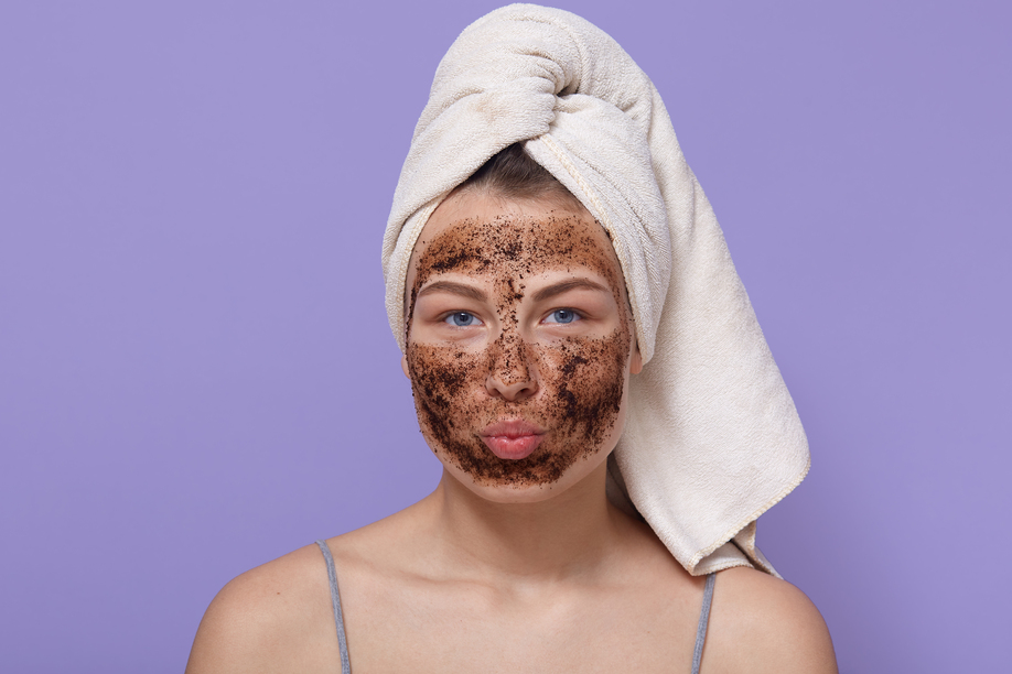 portrait of young attractive woman with white towel on her head, posing isolated over lilac background with pouty lips, looks directly at camera, having chocolate scrub or mask for moisturising.