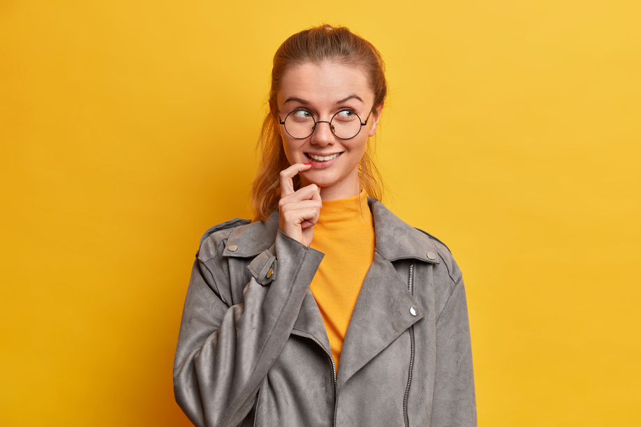 Studio shot of pleasant looking mysterious millennial woman keeps finger near mouth looks away with intention in mind makes planning dressed in grey fashionable jacket isolated on yellow background