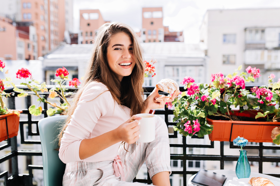 Portrait pretty girl  having breakfast on balcony surround flowers in the sunny morning in city. She holds a cup, croissant, smiling to camera