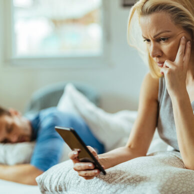 Suspicions woman using boyfriend's mobile phone while he is sleeping on the bed behind her.