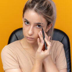 looking at camera young beautiful girl sits at table with makeup tools draw arrow with eyeliner isolated on orange background